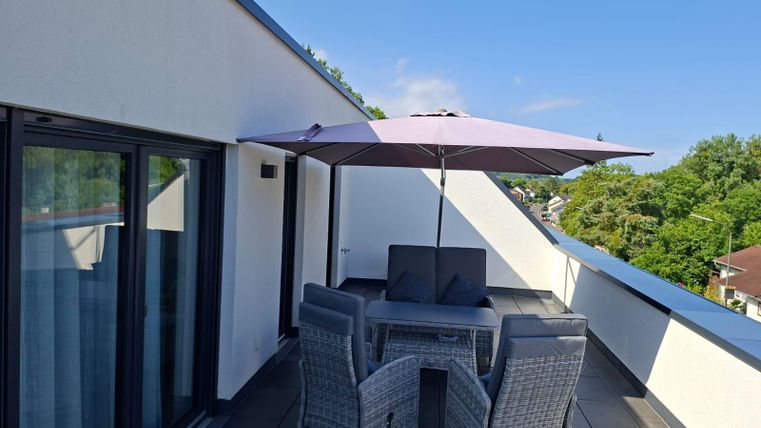  une terrasse moderne avec une table à manger et des chaises. Un parasol fournit de l'ombre, entouré d'arbres verts et d'un ciel dégagé.