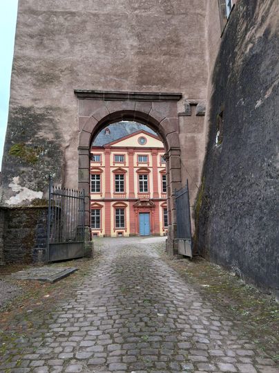 Ein historischer Durchgang mit einem bogenförmigen Tor. Im Hintergrund ist ein malerisches Gebäude mit rosa Fassade und blauen Fenstern zu sehen.
