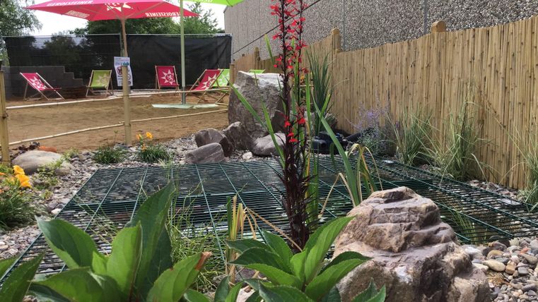 Ein Gartenbereich mit Steinen und Pflanzen, umgeben von einem Holzzaun. Zwei Sonne-Schirme in Grün und Rot bieten Schatten und eine entspannte Atmosphäre.