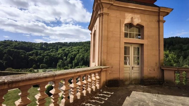 A historical building with an open terrace and large windows. In the background, a forest stretches out under a clear blue sky.