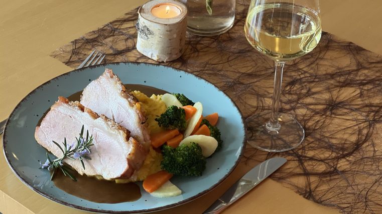 Ein Teller mit saftigem Fleisch, Gemüse und Kartoffelpüree. Daneben steht ein Glas Weißwein auf einem eleganten Tisch.