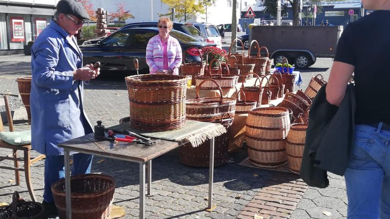 A market stand with handmade baskets displayed on a table. A man is working on the baskets while a woman watches nearby.
