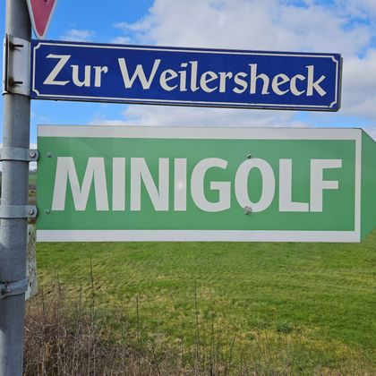 Ein Wegweiser zeigt auf die Minigolfbahn. Im Hintergrund ist eine grüne Wiese unter einem blauen Himmel zu sehen.