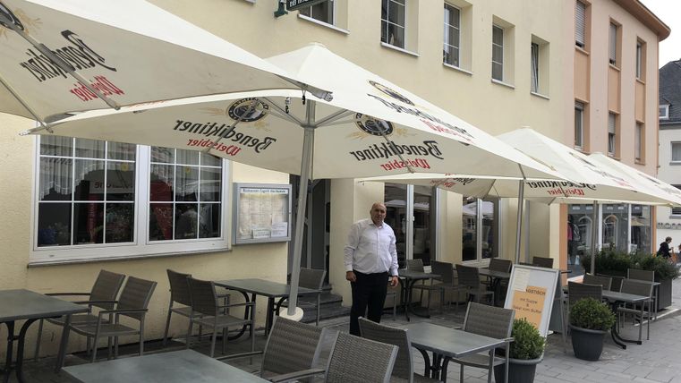 Un restaurant accueillant avec une terrasse extérieure et des parasols. Un serveur est prêt à accueillir les clients.