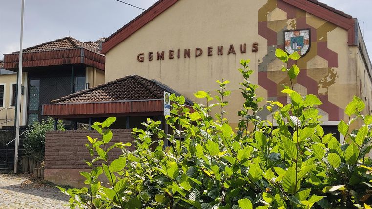 Ein Gemeindehaus mit einem modernen Design und einem Wappen an der Fassade. Im Vordergrund sind grüne Pflanzen zu sehen.