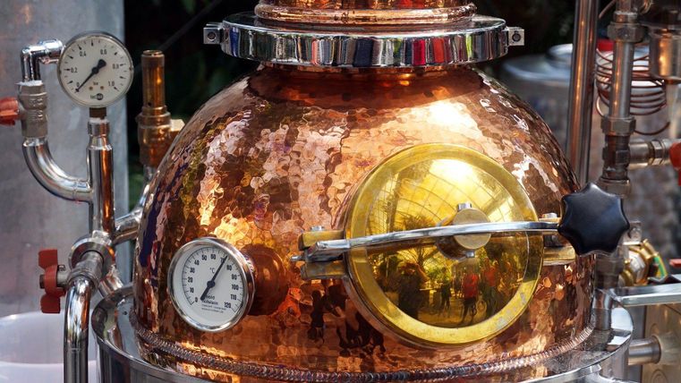 Eine Kupferdestille mit glänzender Oberfläche und verschiedenen Druckmessern. Sie ist Teil eines Destillationsprozesses zur Herstellung von Spirituosen.