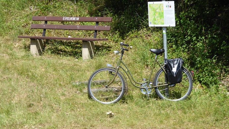 Fahrrad neben einer Bank mit der Aufschrift 'Eifelverein' und einem Wegweiser im Grünen.