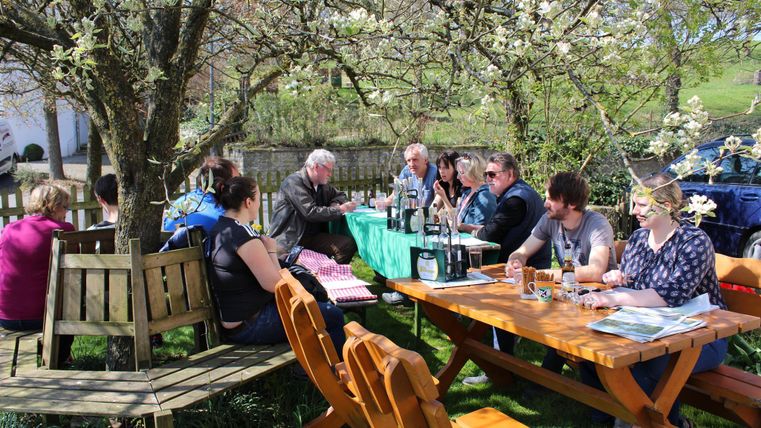 Eine Gruppe von Menschen sitzt an Tischen im Freien unter einem blühenden Baum. Es scheint ein sonniger Tag zu sein, ideal für ein Treffen im Freien.