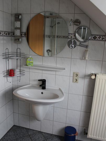 Ein kleines Badezimmer mit einem Waschbecken, einem Spiegel und einem Handtuchhalter. Die Wände sind gefliest, und es gibt eine Heizung sowie einige Regale.