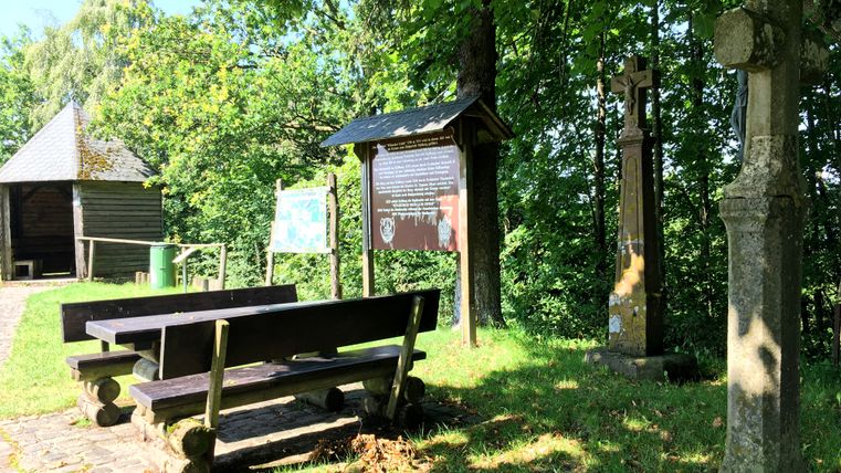 Rastplatz mit Holzbänken, Infotafel und Steinkreuz unter Bäumen.