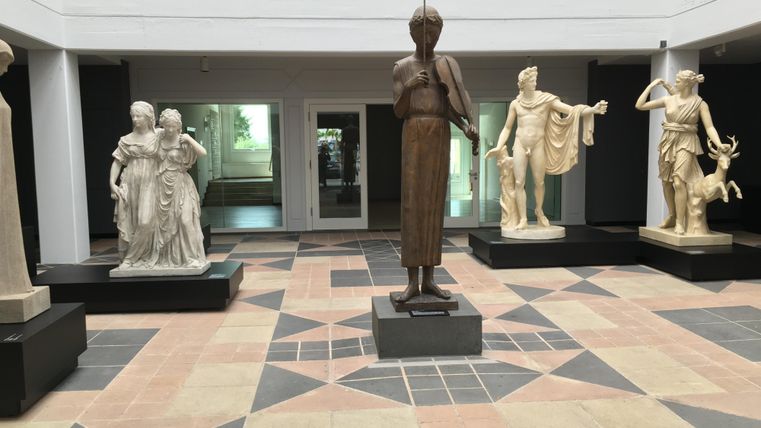 An interior space with several sculptures made from various materials. At the center stands a wooden figure, surrounded by marble statues.