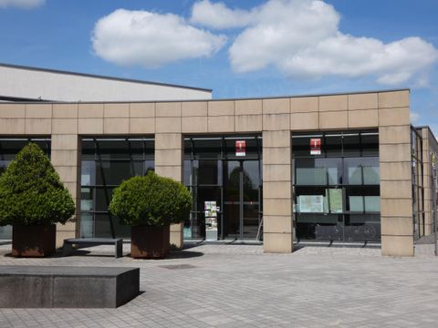 Un bâtiment moderne avec de grandes fenêtres en verre et une architecture contemporaine. Devant le bâtiment se trouvent des plantes bien entretenues et une surface pavée.
