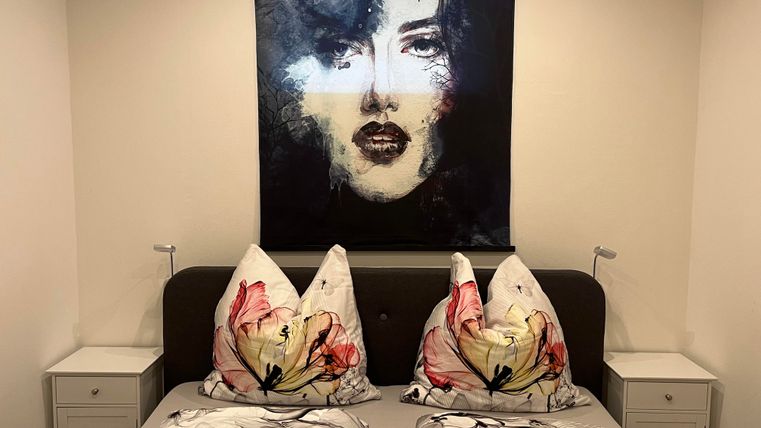 Een moderne slaapkamer met een bed en kleurrijke kussenslopen. Aan de muur hangt een artistiek portret van een vrouw.