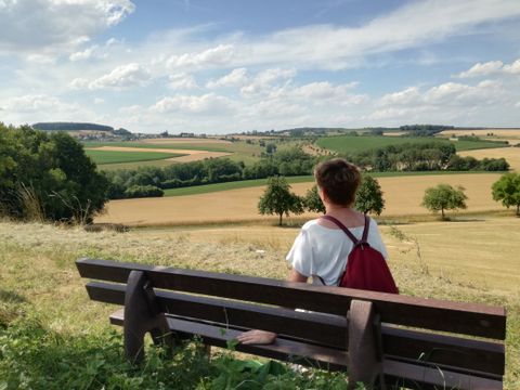 Person sitzt auf einer Bank und blickt auf eine ländliche Landschaft mit Feldern und Bäumen.