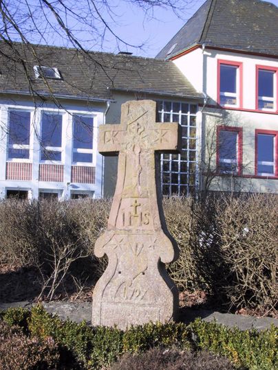 Ein steinernes Kreuz steht im Vordergrund, umgeben von Sträuchern. Im Hintergrund ist ein Gebäude mit großen Fenstern und bunten Rahmen sichtbar.