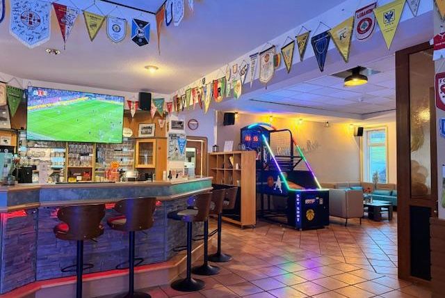 Ein gemütlicher Innenraum eines Lokals mit einer Bar, Sitzgelegenheiten und einer großen Fernsehlenwand für Sportübertragungen. An den Wänden hängen zahlreiche Fahnen und es gibt einen Spielautomat in der Ecke.