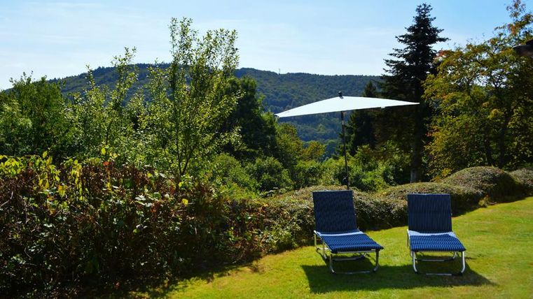 Eine schöne Gartenlandschaft mit zwei Liegen und einem Sonnenschirm. Im Hintergrund sind sanfte Hügel und Bäume zu sehen.
