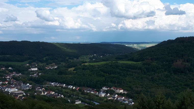 Eine weite Aussicht auf grüne Hügel und ein kleines Dorf. Der Himmel ist bewölkt mit einigen blauen Stellen.