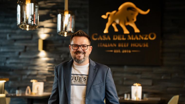 Ein lächelnder Mann steht in einem eleganten Restaurant mit der Beschriftung "Casa del Manzo" im Hintergrund. Die Atmosphäre ist modern und einladend.