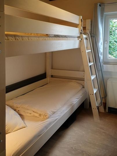 Ein einfaches Zimmer mit einem Etagenbett und einem Fenster. Die Wände sind hell und der Raum ist gut beleuchtet.