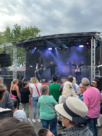 Een band speelt op een podium voor een enthousiast publiek. Veel mensen staan samen en genieten van de muziek onder een bewolkte lucht.