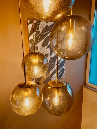 A stylish light design with five round, golden lamps hangs from the ceiling. In the background, there is a decorative picture with modern patterns.