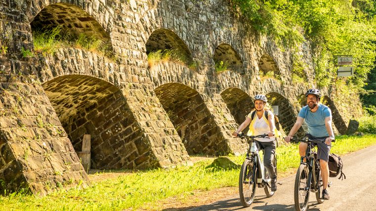 Zwei Radfahrer auf einem Weg neben einer alten Steinmauer mit Bögen, umgeben von grüner Vegetation.