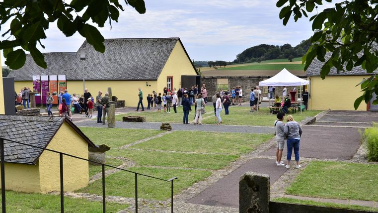 Een groep mensen verzamelt zich op een uitgestrekt terrein met gele gebouwen. Op de achtergrond zijn tenten en een groene landschap zichtbaar.