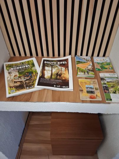 Eine Auswahl an Broschüren und Flyern über die Region Süd-Eifel liegt auf einem Holztisch. Die Materialien informieren über Sehenswürdigkeiten und Aktivitäten in der Umgebung.