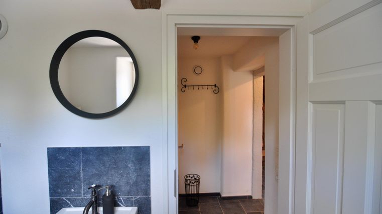 A modern bathroom with a round mirror and an elegant sink installation. In the background, a corridor with a door and hooks is visible.