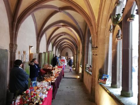 Ein historischer Gang mit gotischen Bögen. An den Wänden stehen Tische mit verschiedenen Handwerksprodukten und Besucher erkunden die Stände.