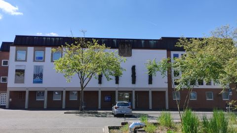 A modern building with multiple windows and a clear sky. In front of the building, there are some trees, and a parking lot is visible.