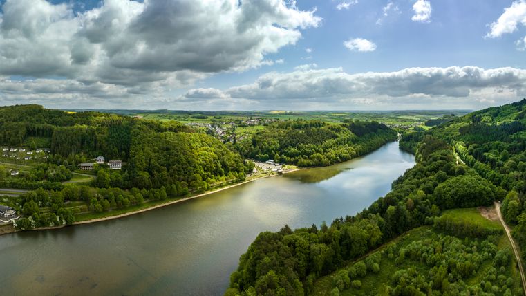 Luftaufnahme des Stausees Bitburg in Biersdorf am See, umgeben von grünen Wäldern und Hügeln unter bewölktem Himmel.
