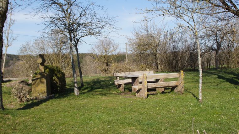 Ein Rastplatz mit Holzbänken und einem steinernen Kreuz auf einer Wiese, umgeben von Bäumen.