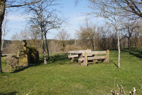 Ein Rastplatz mit Holzbänken und einem steinernen Kreuz auf einer Wiese, umgeben von Bäumen.