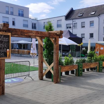 Une entrée accueillante avec une structure en bois et des plantes. En arrière-plan, on peut voir des tentes et des bâtiments modernes.