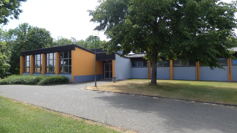 Ein modernes Gebäude mit orangefarbenen und grauen Wänden. Vor dem Gebäude ist eine gepflasterte Straße und grüne Bäume.