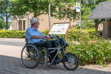Ein Mann im Rollstuhl sitzt liest ein Schild auf einem Rundweg