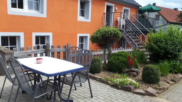Ein helles Wohnhaus mit einer orangefarbenen Fassade. Im Vordergrund steht ein Tisch mit Stühlen und ein gepflegter Garten mit grünen Pflanzen.