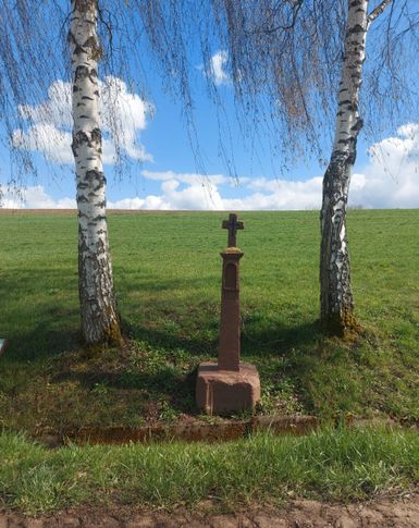 Ein Wegekreuz steht zwischen zwei Bäumen.