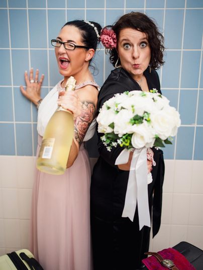 Twee blije vrouwen poseren in een badkamer. De een houdt een fles champagne vast en de ander een boeket bloemen.