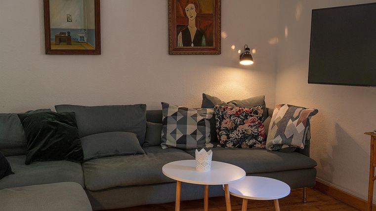 Ein gemütliches Wohnzimmer mit einem grauen Sofa und bunten Kissen. An der Wand hängen zwei Bilder, und es stehen zwei kleine, weiße Tische.