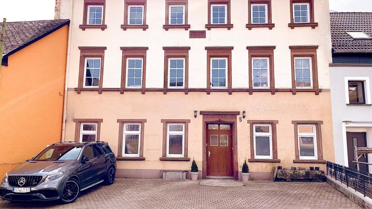 A three-story building with a light façade and brown window frames. In front of the house, there is a black car parked on a paved parking lot.