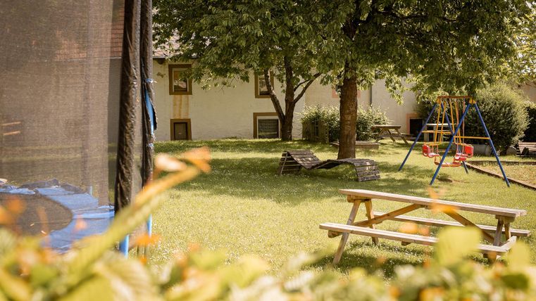 Ein schöner Garten mit einer Wiese, einem großen Baum und einer Schaukel. Auf der linken Seite steht ein Trampolin, und im Hintergrund sieht man ein Haus.