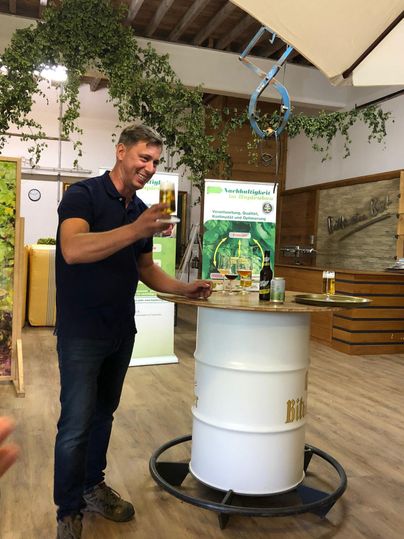 A man is holding a glass with a drink and is standing at a high table. In the background, decorative elements and a bar are visible.