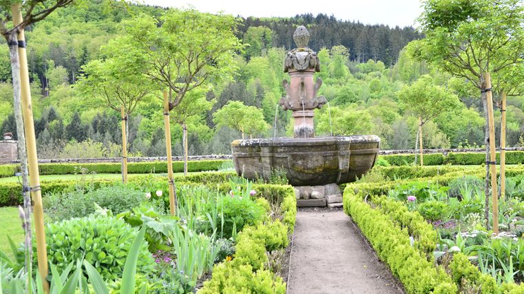 Ein schöner Garten mit einem Brunnen in der Mitte. Umgeben von grünen Pflanzen und Bäumen.