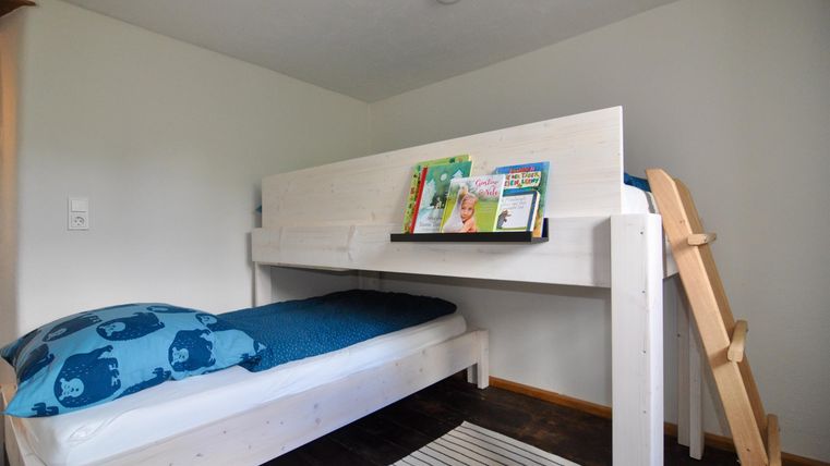 A cozy loft bed in a modern room. A blue blanket lies on the bed and a shelf with books is visible.