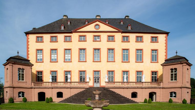An impressive historic building with an elegant pink facade and large windows. The expansive lawn and the staircase give the castle a magnificent impression.