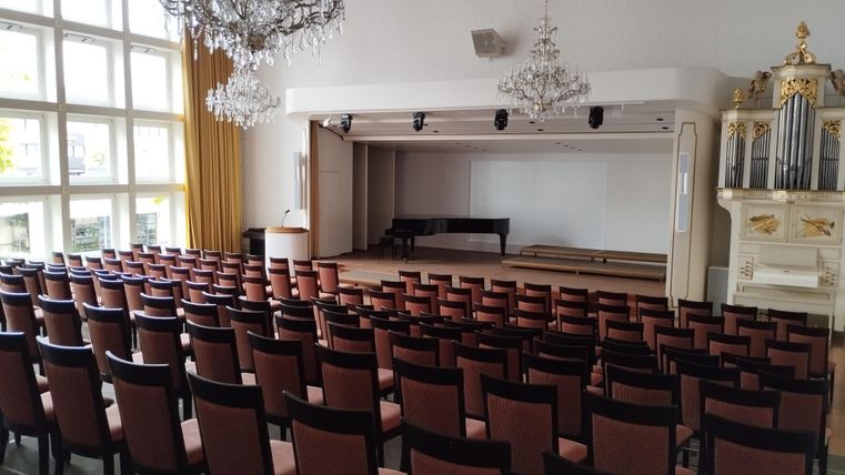 Een elegante evenementenruimte met veel stoelen en een piano op het podium. Grote ramen zorgen voor veel licht in de ruimte.