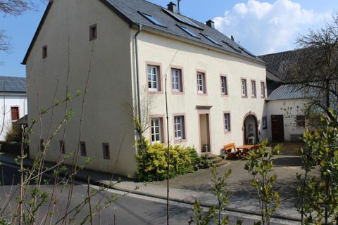 Ein charmantes Haus mit hellem Putz und Fensterrahmen in Pastellfarben. Im Vordergrund befinden sich Pflanzen und eine ruhige Straße.
